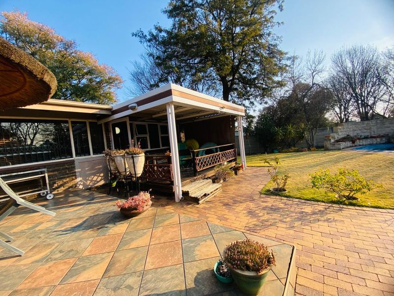 A spacious outdoor entertainment area featuring a covered patio with timber decking, adjacent paved terrace, and access to a garden and swimming pool.
