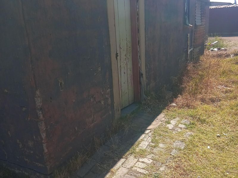 Exterior view of the rear building facade showing significant wear to the wall finishes and door frame, with adjacent brick paving and overgrown grass.