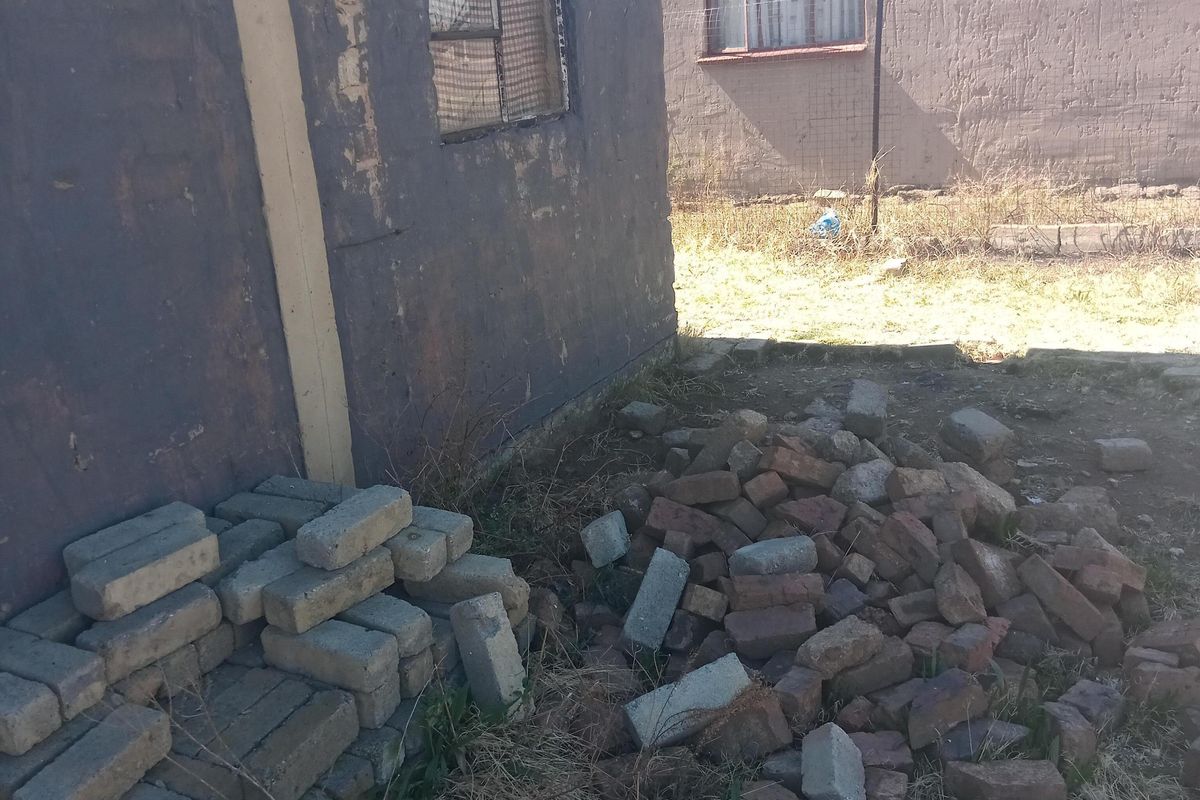 Rear exterior view of a residential property showing weathered masonry walls and security window grilles with loose building materials in the yard.