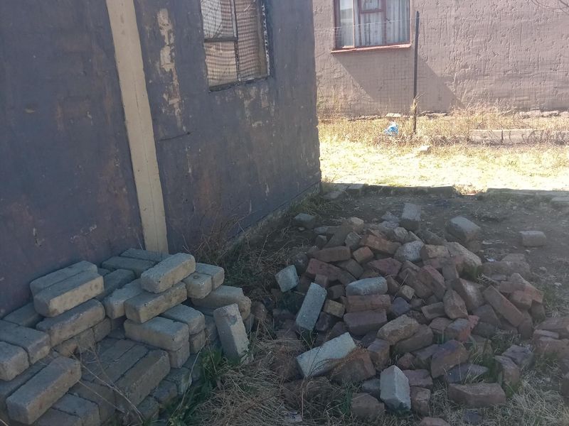 Rear exterior view of a residential property showing weathered masonry walls and security window grilles with loose building materials in the yard.