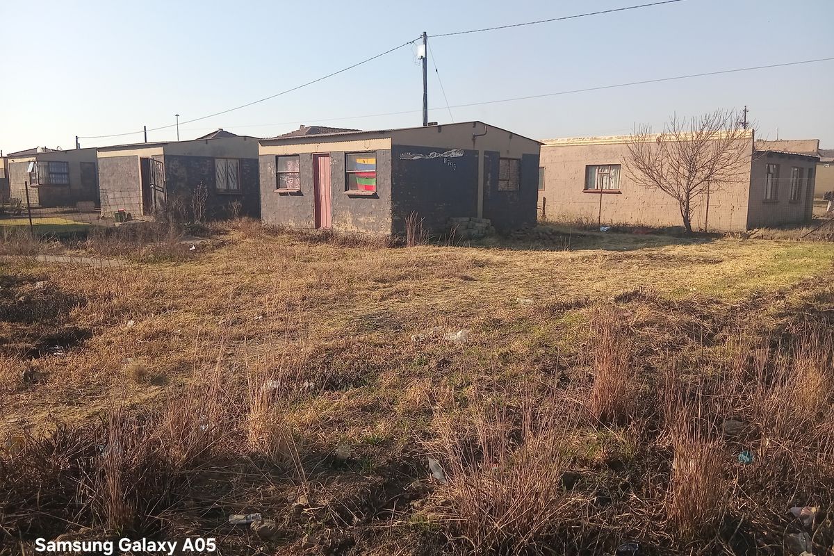 Exterior view of a detached residential property featuring masonry construction and a corrugated metal roof, set on a large plot with overgrown grounds.