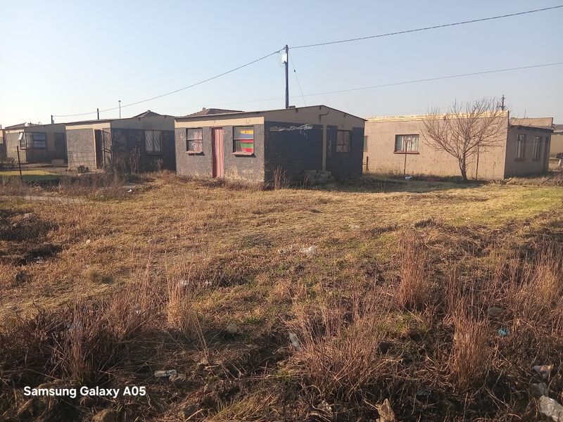 Exterior view of a detached residential property featuring masonry construction and a corrugated metal roof, set on a large plot with overgrown grounds.
