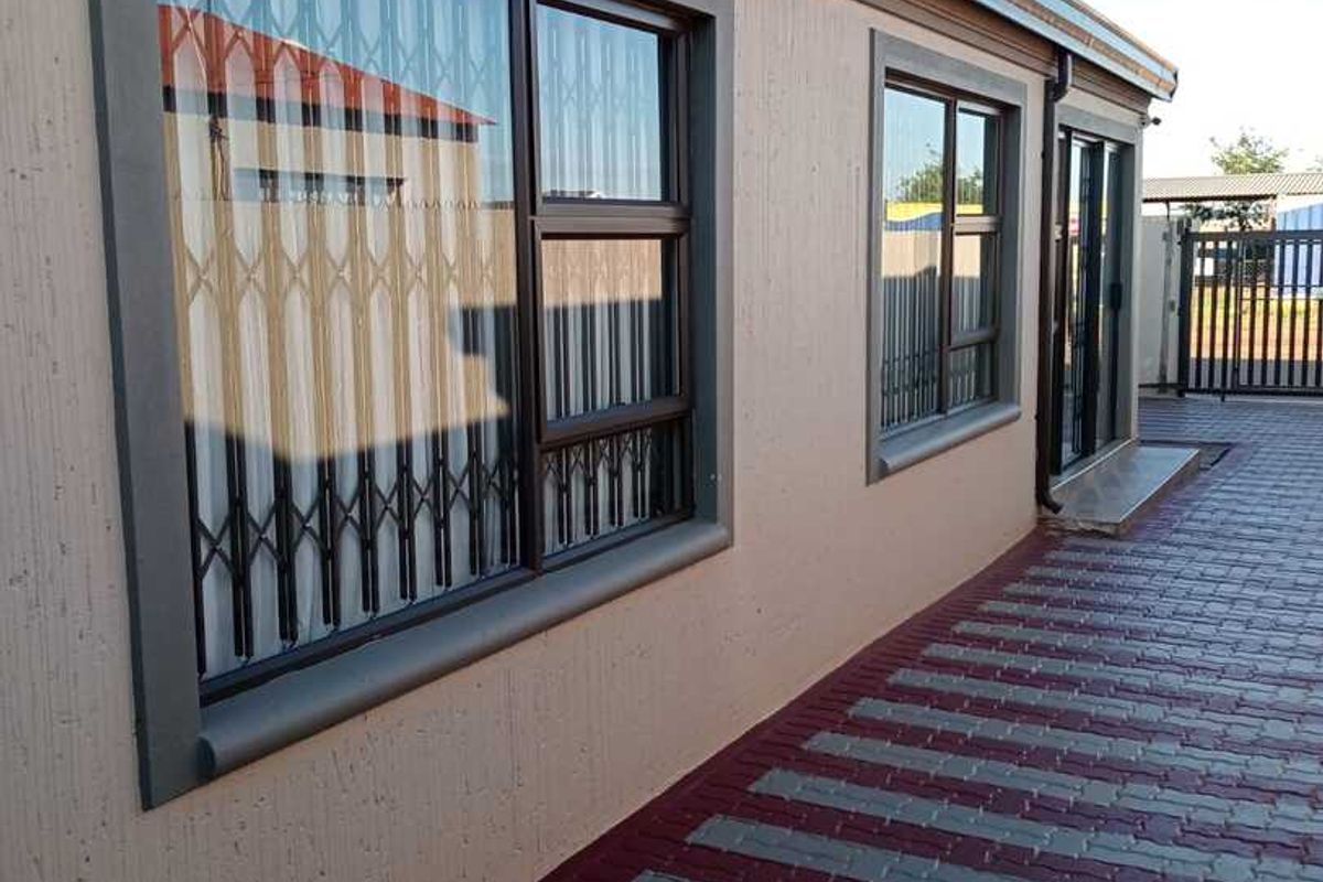 A well-maintained rear exterior view featuring paved ground, aluminium window frames with security gates, and a sliding door entrance.