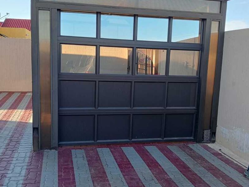 A modern garage structure featuring a dark-framed sectional door with glass panels and a distinctive arched polycarbonate roof, set on a paved driveway.
