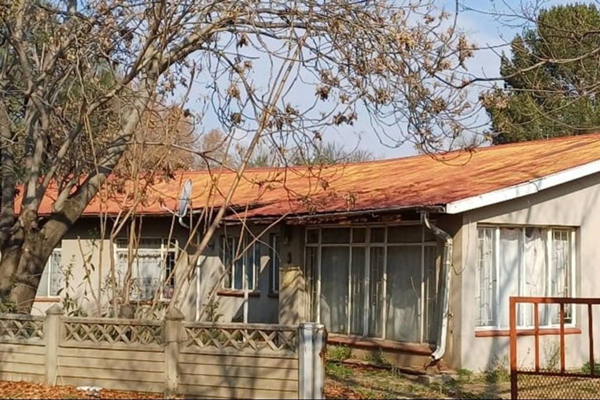 Front elevation of a single-story house featuring a metal roof, precast boundary wall, and metal gate, showing signs of significant exterior wear.