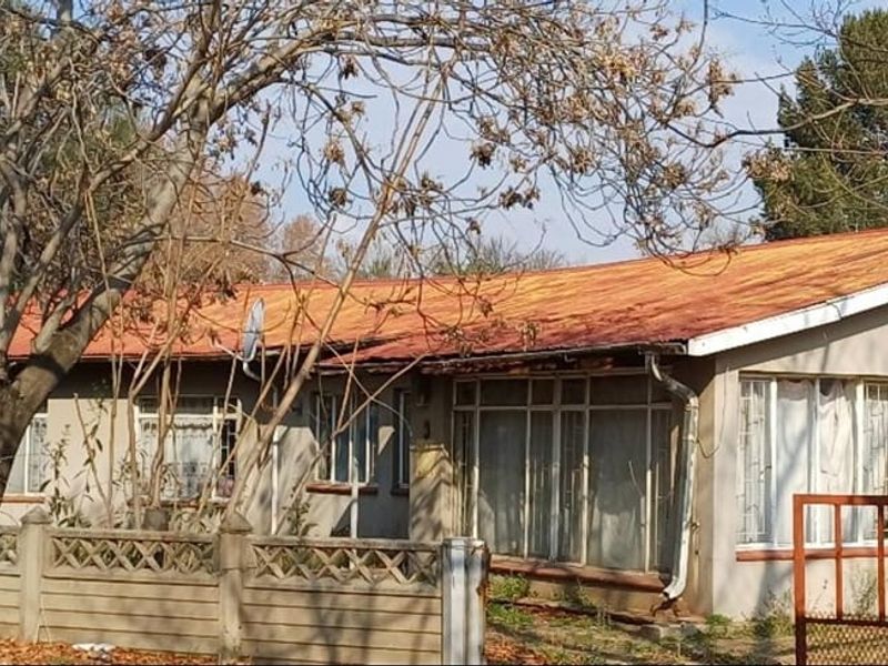 Front elevation of a single-story house featuring a metal roof, precast boundary wall, and metal gate, showing signs of significant exterior wear.