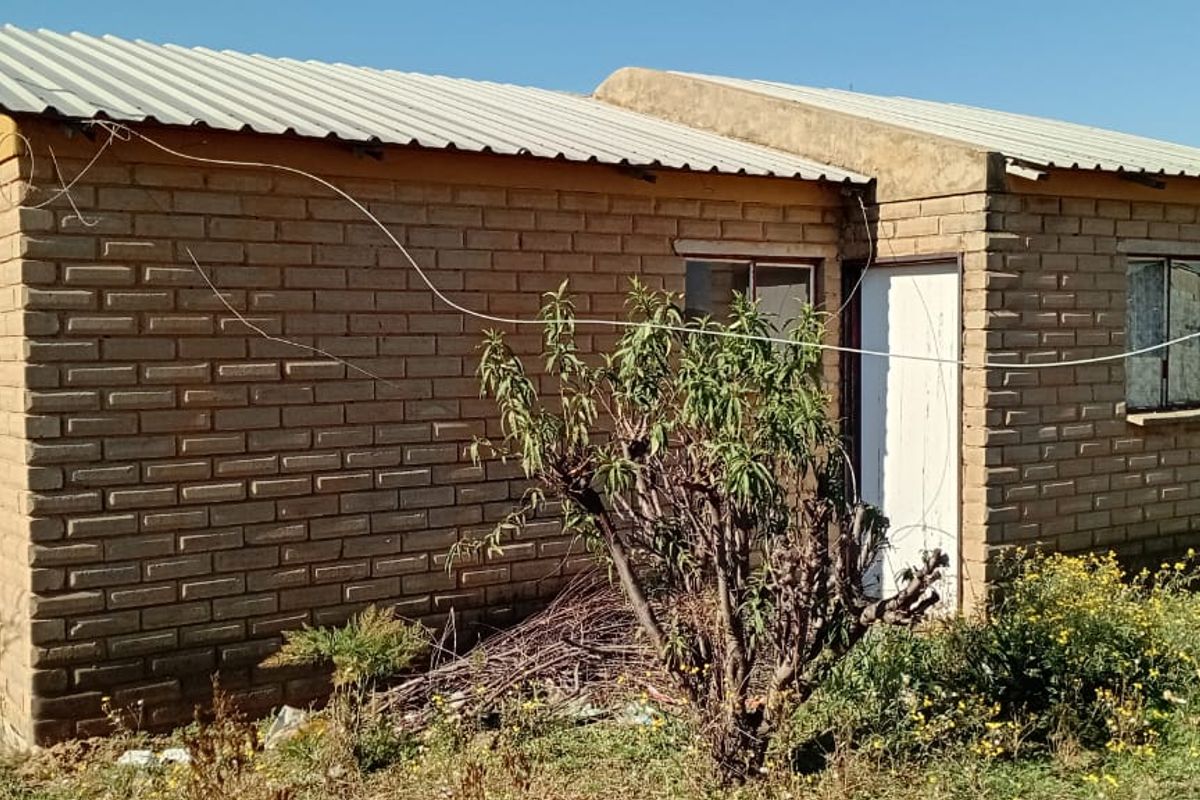 Front view of a single-story detached house featuring exposed brick walls and a corrugated metal roof, situated on an unlandscaped plot.