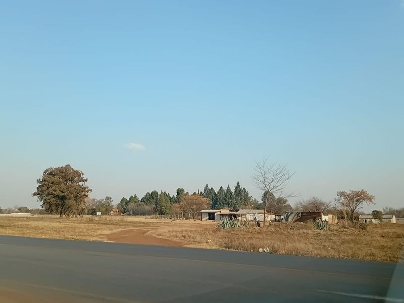 A view of the small holding property from the adjacent asphalt road, showing the open land and existing outbuildings.