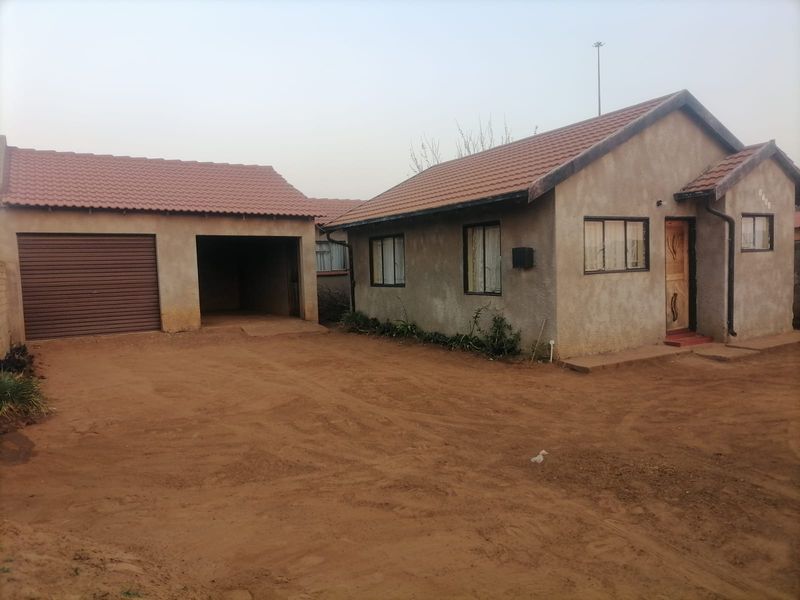 Front view of a single-story house featuring a tiled roof, a garage, and an open carport area with an unpaved driveway.