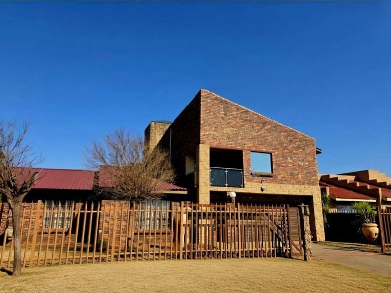 Front elevation of a double-story face brick house featuring a balcony with glass balustrades, metal roofing, and secure perimeter fencing.