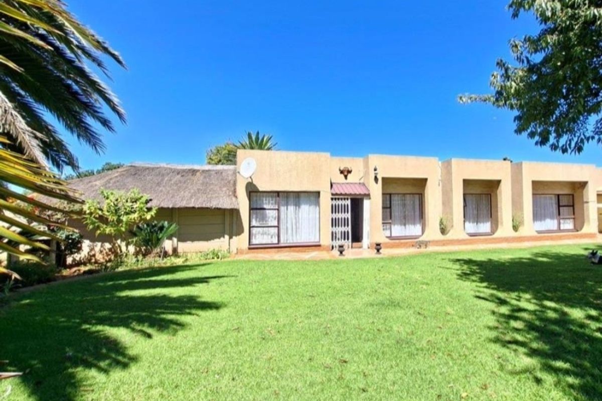 Rear view of a single-story house featuring a thatched roof section, stucco facade, and large windows overlooking a spacious green lawn.
