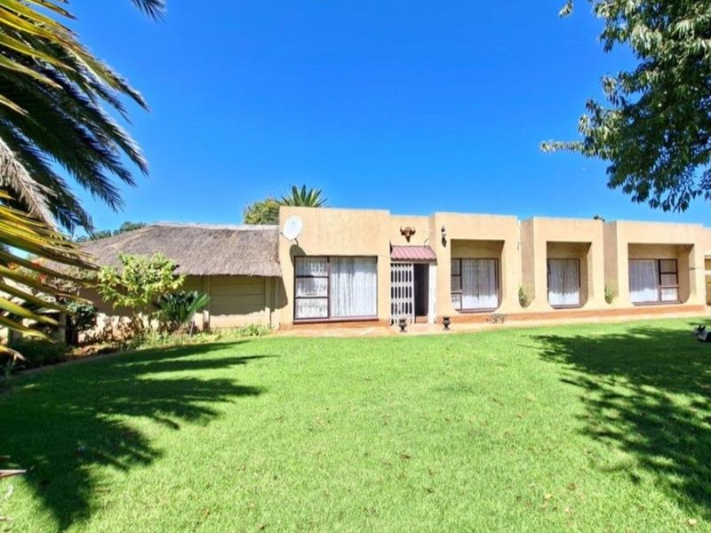 Rear view of a single-story house featuring a thatched roof section, stucco facade, and large windows overlooking a spacious green lawn.