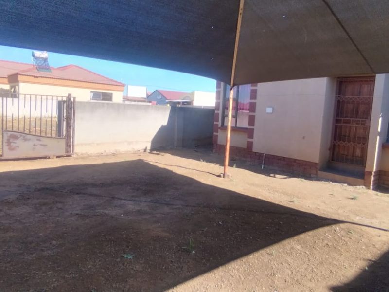 Rear exterior area featuring a shade sail, perimeter wall, and metal gate access, with an unpaved yard space.