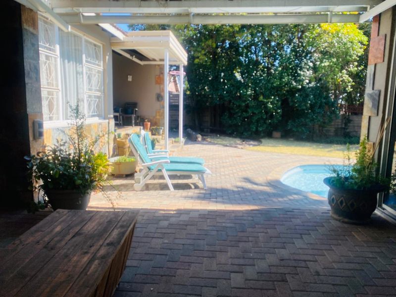 A covered paved patio area overlooking a swimming pool and garden, featuring stone wall accents and a sheltered outdoor living space.