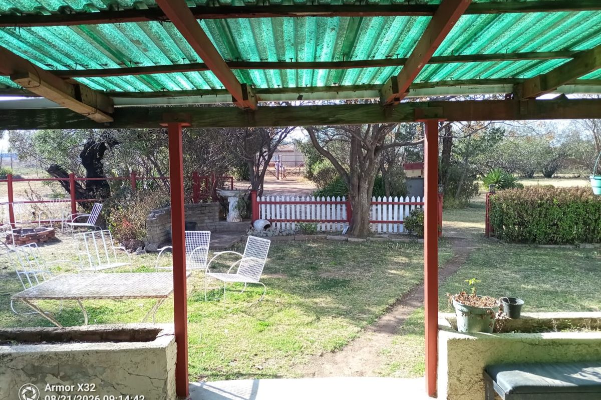 Covered outdoor patio area featuring a corrugated roof, timber support structure, and built-in concrete planter boxes overlooking a garden with a picket fence.