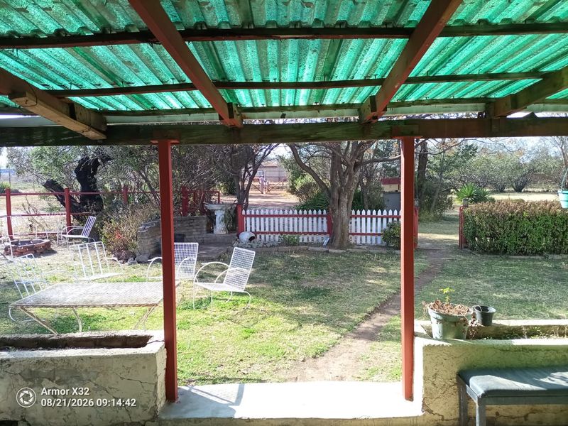 Covered outdoor patio area featuring a corrugated roof, timber support structure, and built-in concrete planter boxes overlooking a garden with a picket fence.