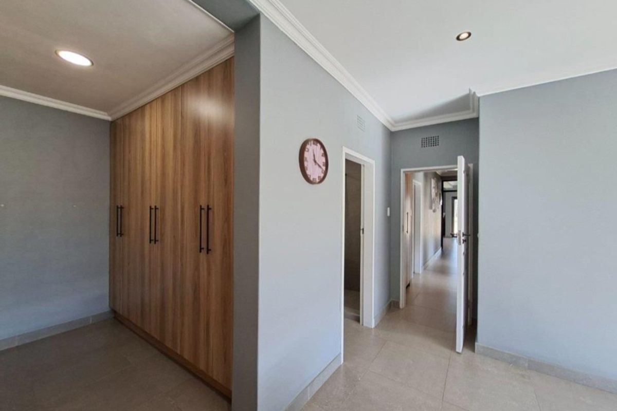 Spacious bedroom featuring modern wood-finish built-in cupboards, neutral tiled flooring, recessed downlights, and access to an adjoining hallway.