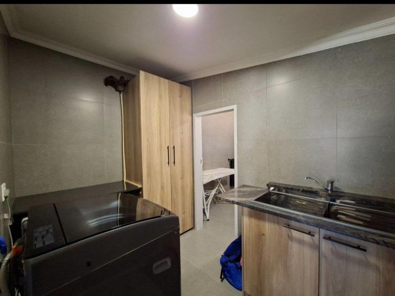 A functional laundry room featuring modern wood-finish cabinetry, stone countertops, a stainless steel sink, and neutral wall tiling.