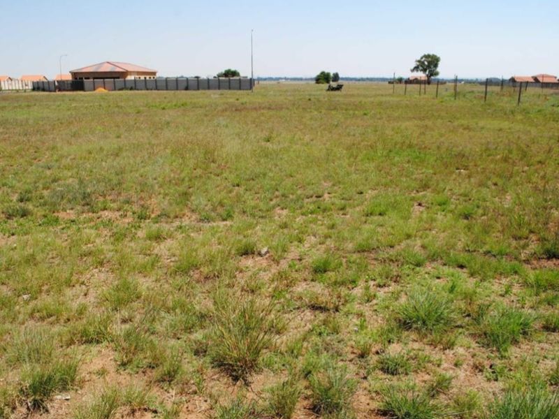 A wide, level vacant residential plot featuring natural grass cover and perimeter wire fencing, offering a clear space for future development.