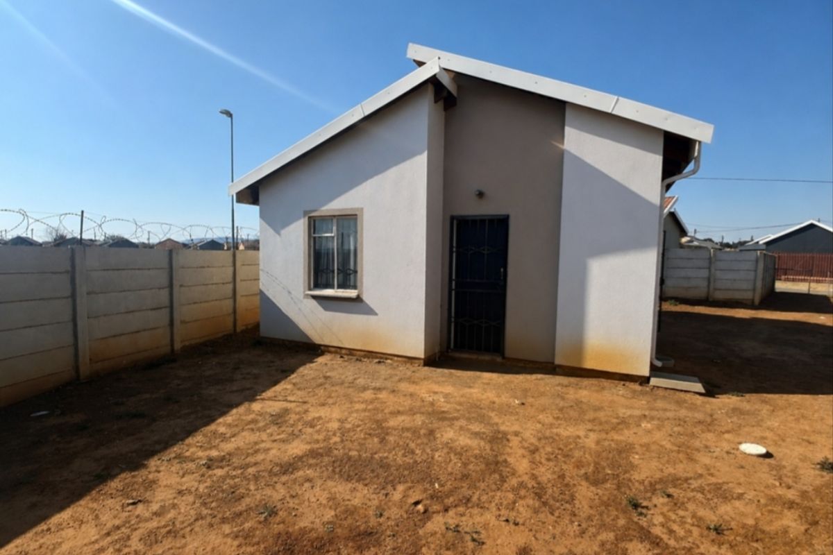 Rear view of a detached house featuring a simple facade, security gate, and a large, unlandscaped yard enclosed by a precast concrete boundary wall.