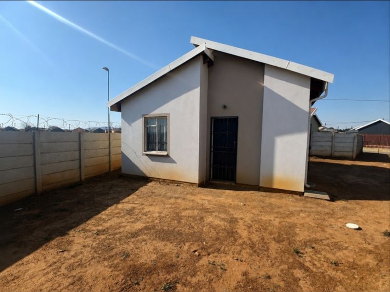 Rear view of a detached house featuring a simple facade, security gate, and a large, unlandscaped yard enclosed by a precast concrete boundary wall.