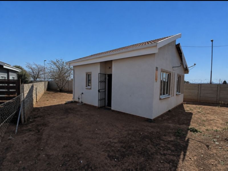 Rear view of a detached house featuring a tiled roof, security gate, and a large, unlandscaped yard enclosed by precast concrete boundary walls.