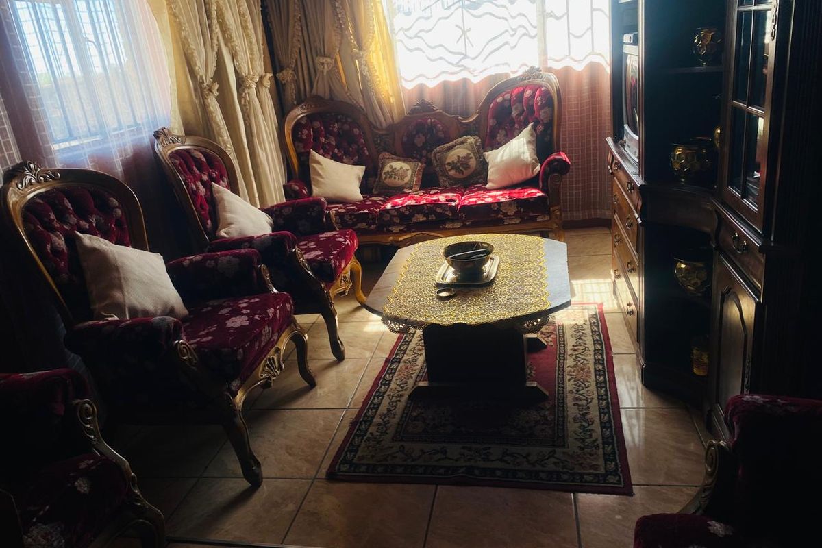 A living room featuring tiled flooring and large windows with security bars, currently furnished with traditional seating.