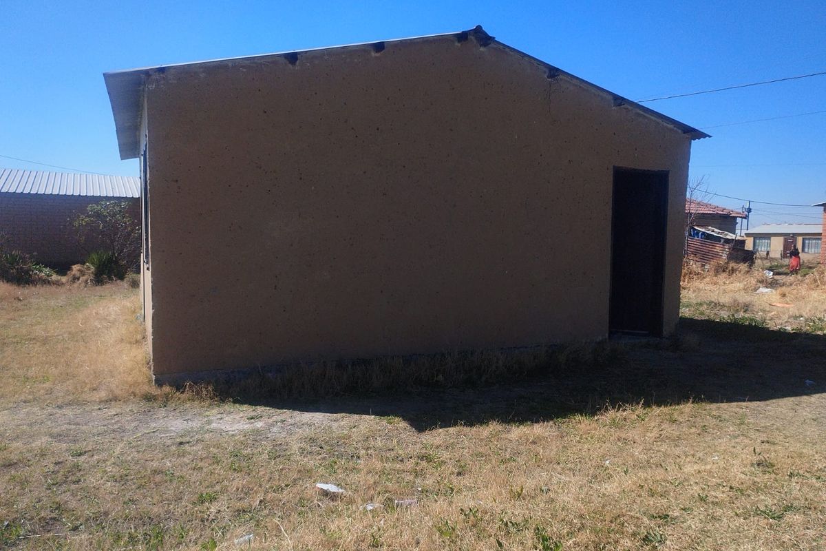 A view of the rear exterior of a simple residential structure with plastered walls and a metal roof set on a dry grass plot.