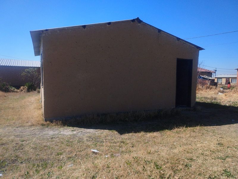 A view of the rear exterior of a simple residential structure with plastered walls and a metal roof set on a dry grass plot.