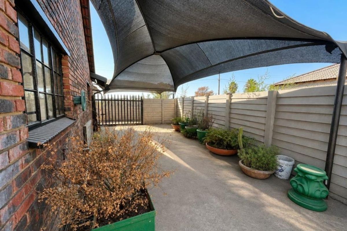 A paved outdoor patio area featuring a large overhead shade sail, enclosed by a precast concrete boundary wall and a metal security gate.
