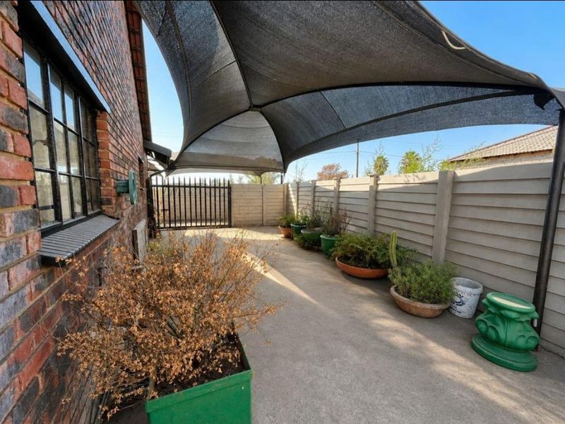 A paved outdoor patio area featuring a large overhead shade sail, enclosed by a precast concrete boundary wall and a metal security gate.