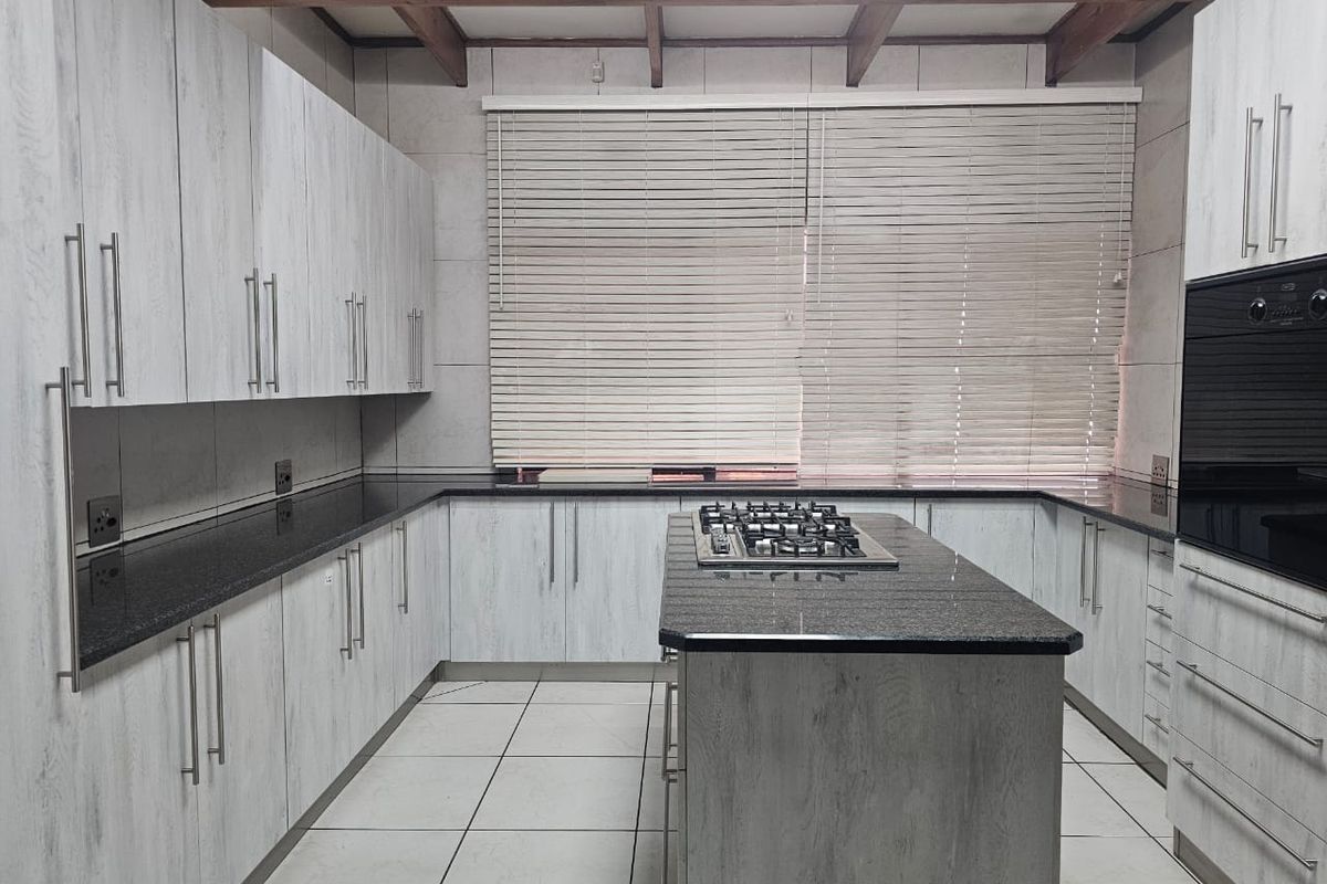 Modern kitchen featuring light wood-grain cabinetry, dark granite countertops, a central island with a gas hob, built-in oven, and exposed timber ceiling beams.