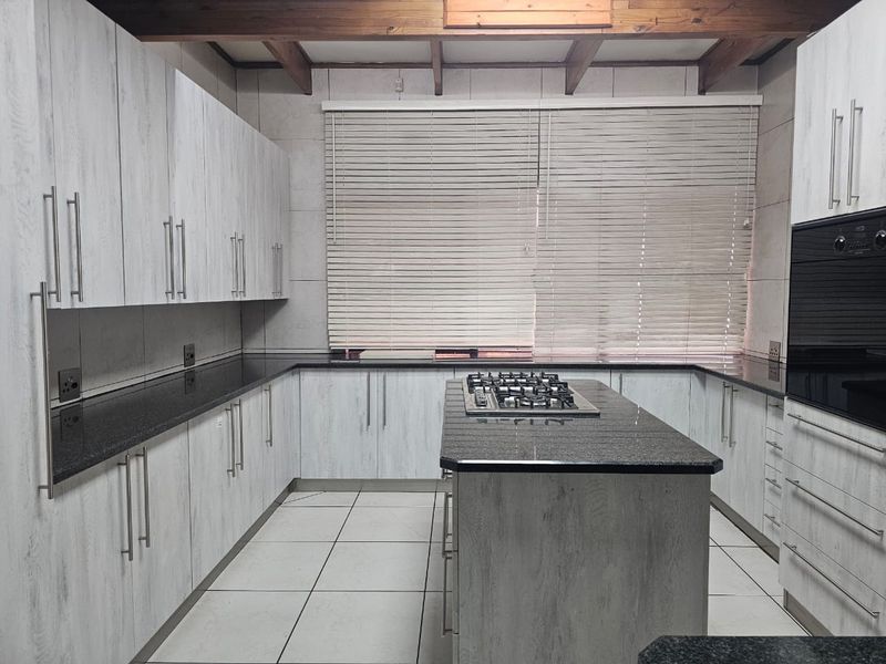 Modern kitchen featuring light wood-grain cabinetry, dark granite countertops, a central island with a gas hob, built-in oven, and exposed timber ceiling beams.