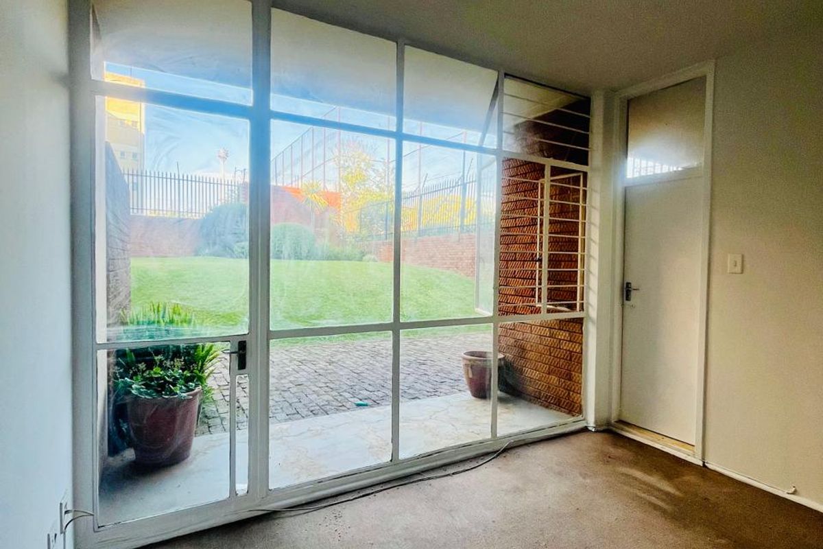 A sunlit living area featuring large floor-to-ceiling windows with direct access to a paved patio and garden space.