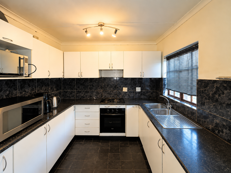 A functional kitchen featuring white cabinetry, dark tiled backsplash, built-in oven with gas hob, and a double bowl sink with a window view.