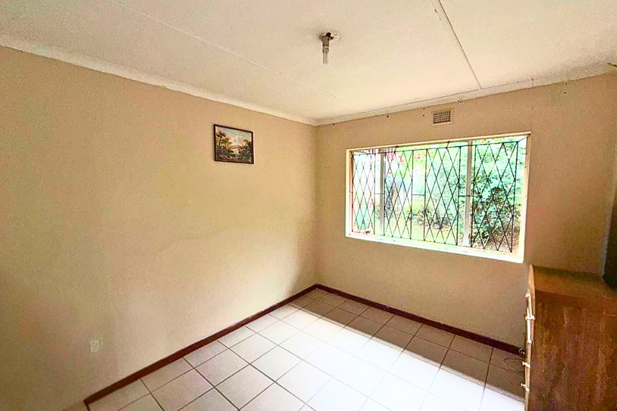 A simple bedroom featuring tiled flooring, a large window with security bars, and neutral wall tones, offering a blank canvas for potential updates.