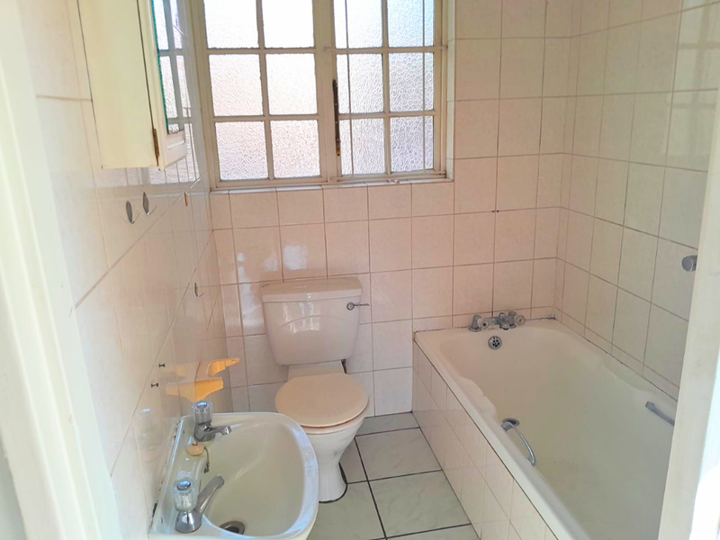 A dated bathroom featuring a built-in bathtub, pedestal washbasin, toilet, and full-height ceramic wall tiling with an obscure glass window.