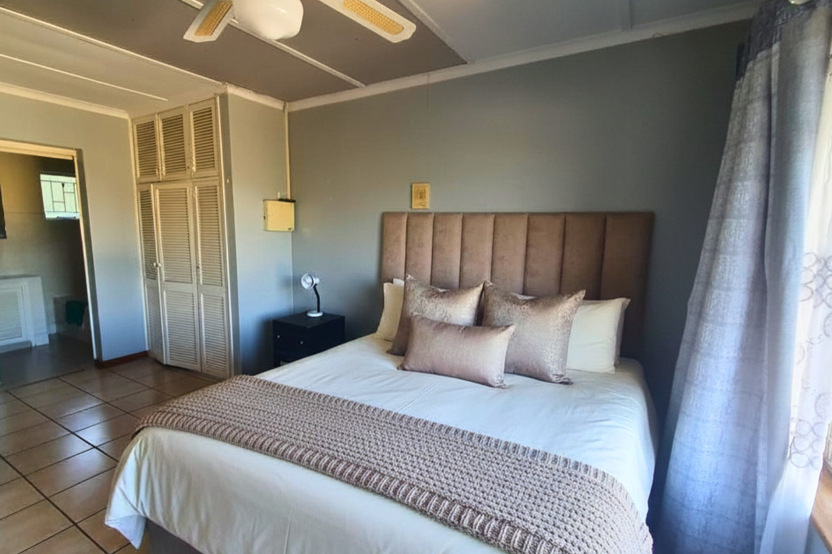 A well-lit bedroom featuring tiled flooring, built-in cupboards, and a ceiling fan, with an adjoining bathroom visible through the doorway.