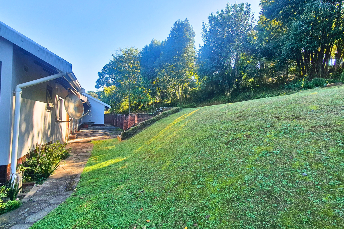 Rear exterior view of the property featuring a stone paved walkway, brick retaining wall, and a sloped garden area with mature trees.
