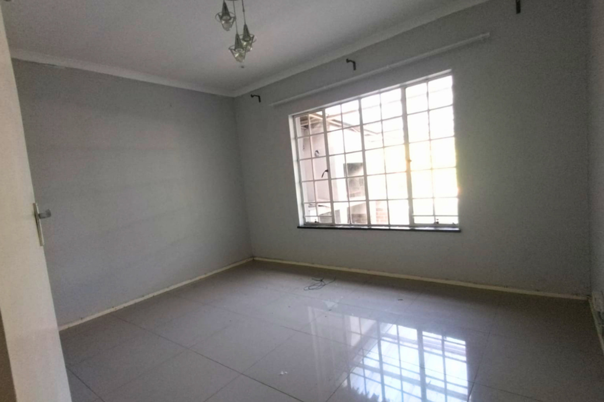 A vacant bedroom featuring neutral grey walls, polished floor tiles, and a large window with security bars, offering a clean and versatile living space.