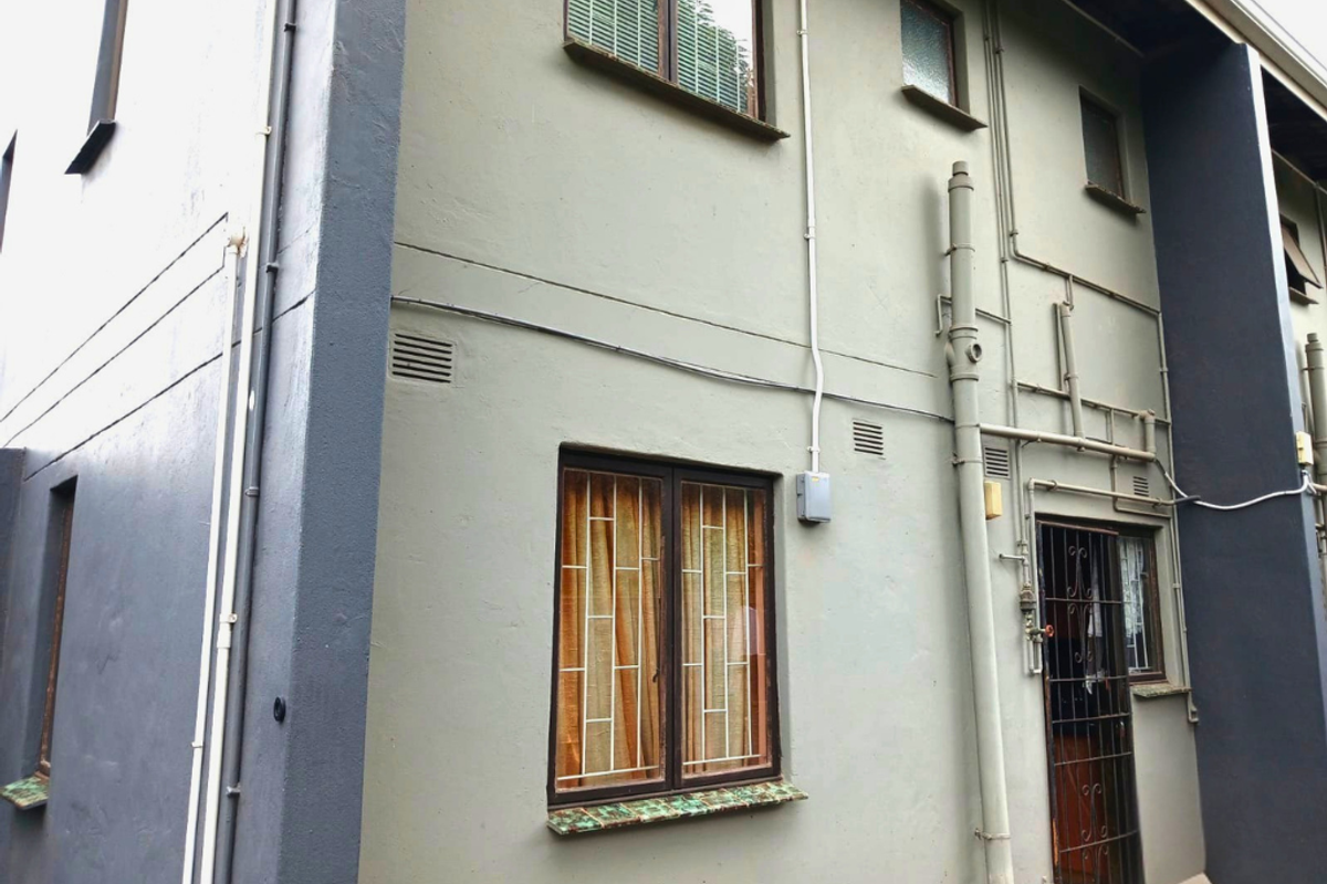 A view of the townhouse exterior facade featuring painted masonry walls, security bars on windows and doors, and exposed external plumbing infrastructure.