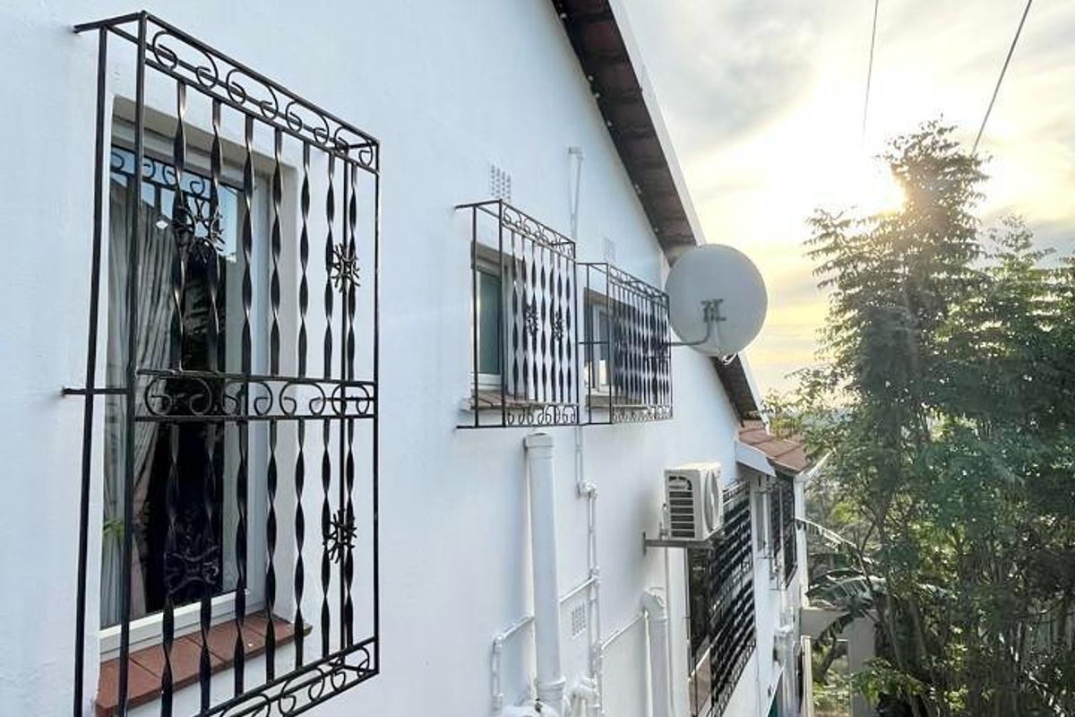 Side exterior view of the property featuring security bars on windows, an external air conditioning unit, and a concrete pathway leading to the rear.