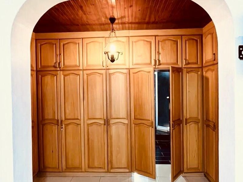 A bedroom featuring extensive built-in wooden cabinetry, a warm timber ceiling, and neutral tiled flooring, accessed through a classic arched doorway.