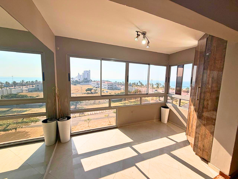 A bright, enclosed balcony featuring large sliding glass windows with expansive coastal views, tiled flooring, and integrated built-in cabinetry.