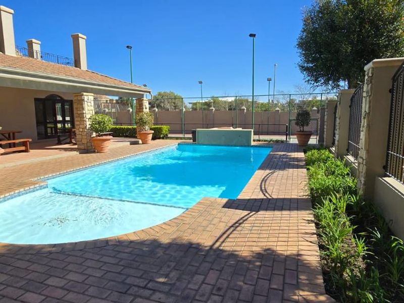 A well-maintained outdoor pool area featuring a swimming pool, brick-paved surround, covered patio with stone-clad pillars, and secure wrought iron perimeter fencing.
