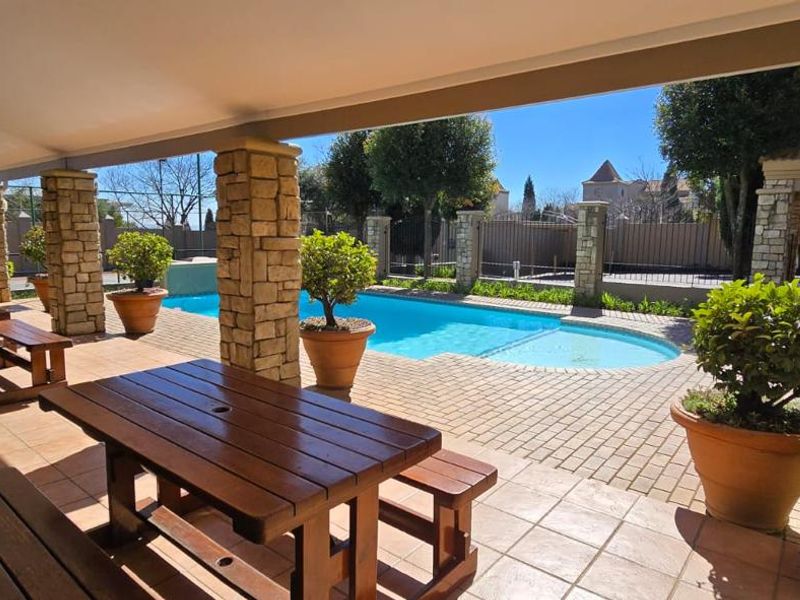 A well-maintained outdoor pool area featuring a covered patio with stone-clad pillars, paved flooring, and direct access to a swimming pool.