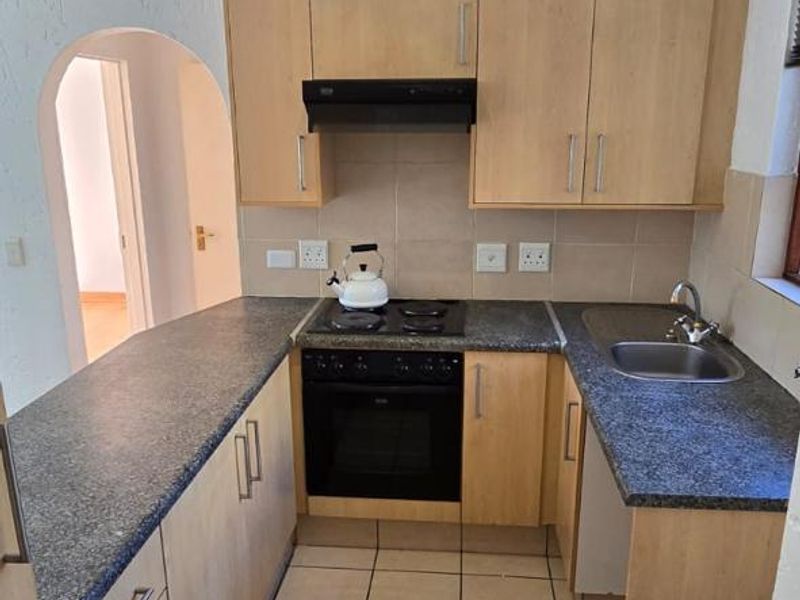 A functional U-shaped kitchen featuring light wood-finish cabinetry, dark laminate countertops, built-in oven, electric hob, and tiled flooring with an adjacent arched doorway.