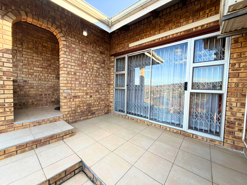 A covered patio area featuring exposed brick walls, tiled flooring, and a large sliding glass door with security gate access.