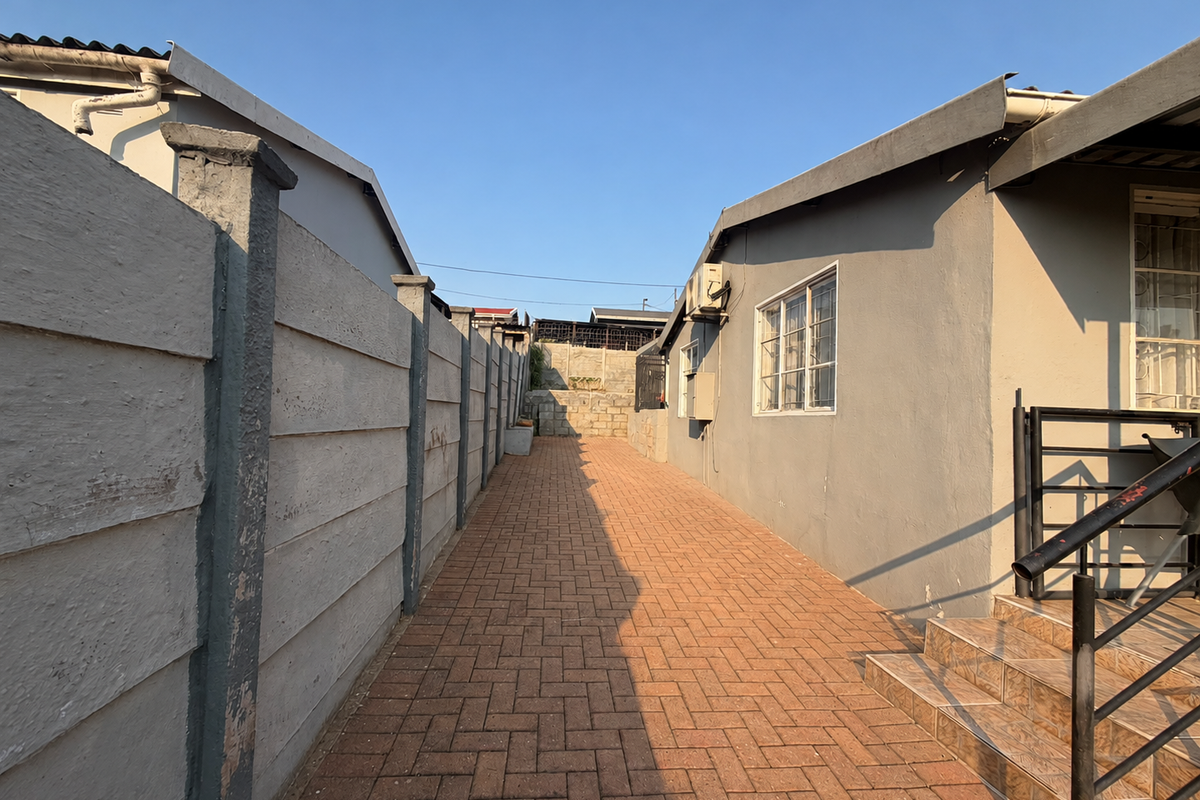 A side exterior view featuring a paved walkway, precast concrete boundary wall, and tiled staircase leading to the side entrance of the house.