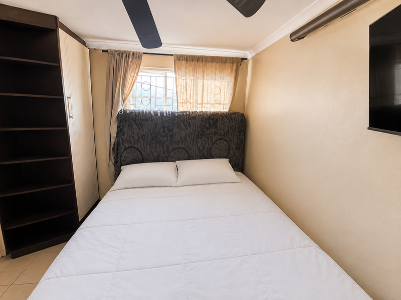 A compact bedroom featuring a built-in shelving unit, ceiling fan, and security bars on the window, providing a functional and secure sleeping space.